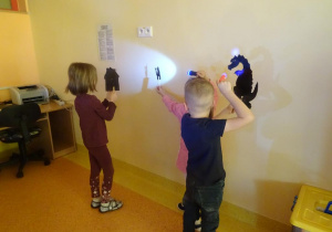 Troje dzieci trzyma w ręku sylwety zwierząt i domku na które święcą latarkami tworząc cień na ścianie.